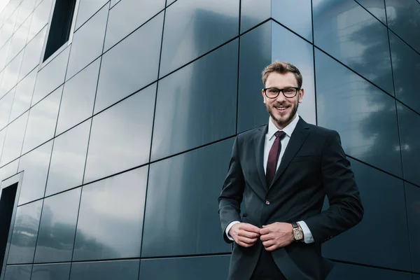 Egy elegáns öltönyös férfi mosolyog egy modern épület előtt.