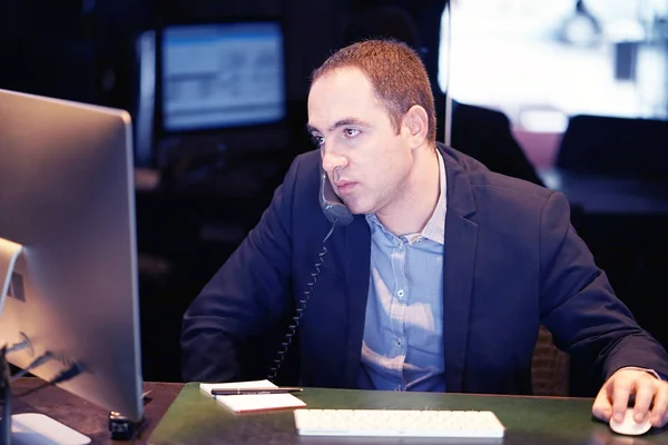 Egy férfi telefonon beszél, miközben számítógép előtt ül, üzleti öltözetben.