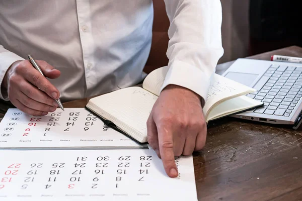 Egy férfi a naptárját nézi, miközben jegyzetel egy laptop mellett.