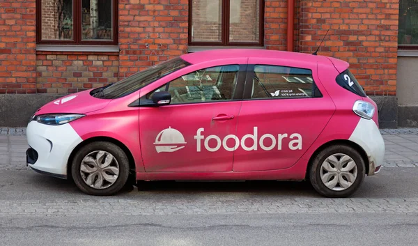 Egy Foodora logóval ellátott rózsaszín autó parkol egy városi utcán.